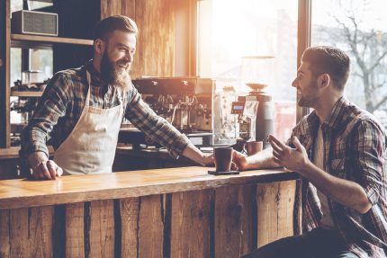 Barista and his customer discussing something with smile while sitting at bar counter at cafe